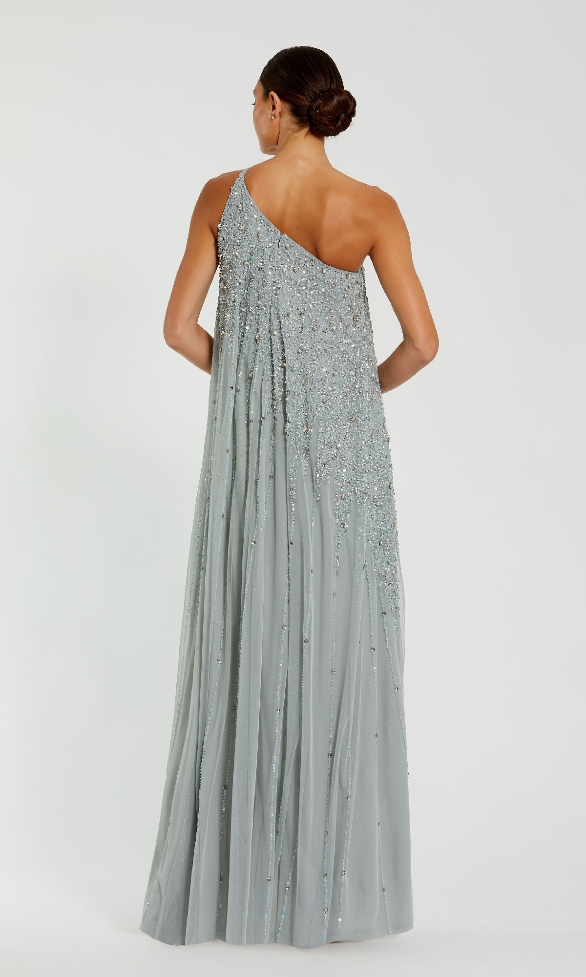 A woman faces away, wearing the Mac Duggal 9224 one-shoulder long formal dress in light gray with bead and sequin embellishments; her hair is styled in a low bun for an elegant look.